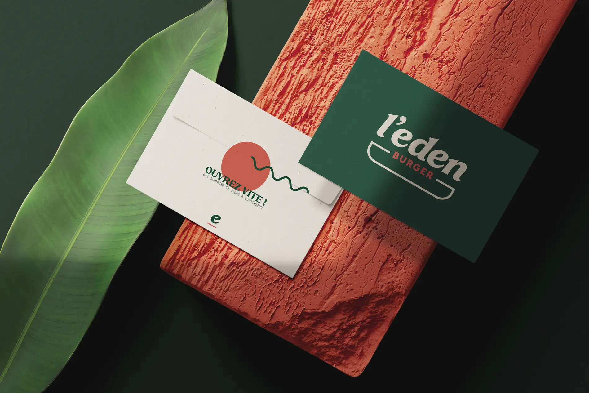 Carte verte au logo L’Eden Burger et enveloppe crème décorée de corail, présentées sur une brique et une feuille.